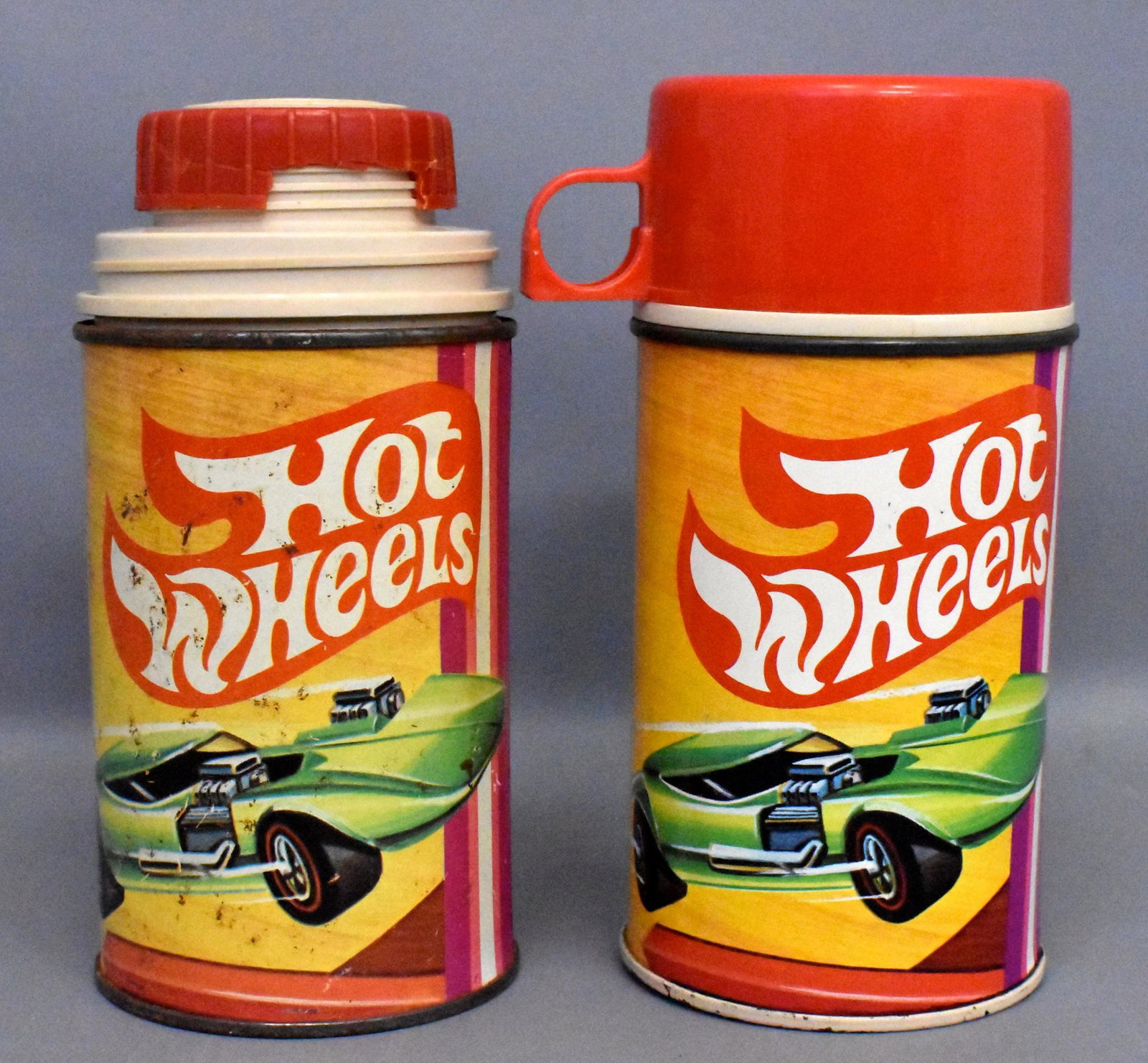 Group of two vintage Hot Wheels Thermoses: One has a tan painted base and is in very good condition complete with tan stopper and cup, the other is in fair condition with light surface rust and unpainted base, it has a red stopper that has a b