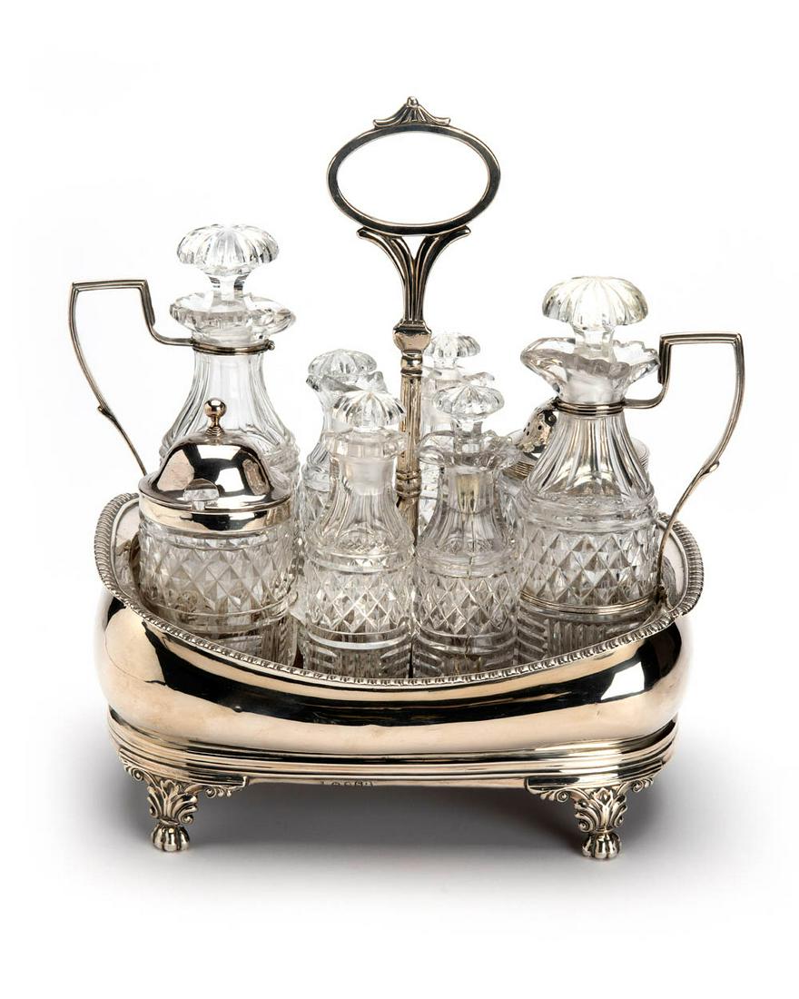 An English silver cruet set: A shaped rectangular model with sloping gadrooned border. On four foliage and claw feet. The holder with six cut glass flasks and two jars with silver covers. Maker Charles Thomas Fox, 1813-1814, Lond
