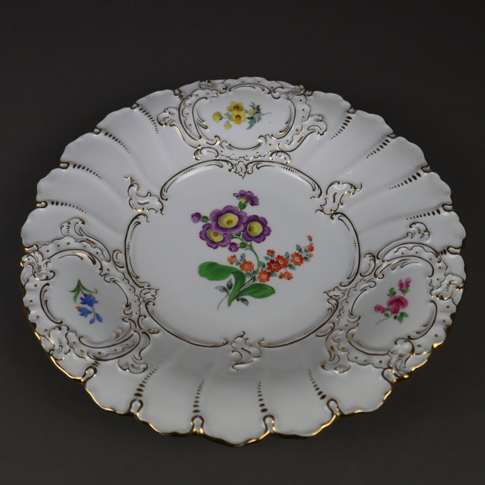 Prunkteller Meissen - unterglasurblaue Schwertermarke: Prunkteller Meissen - unterglasurblaue Schwertermarke mit zwei Schleifstrichen, polychrom und goldbemalt, geschweifter tiefgemuldeter Teller, Fahne dekoriert mit drei reliefierten Rocaillekartuschen m