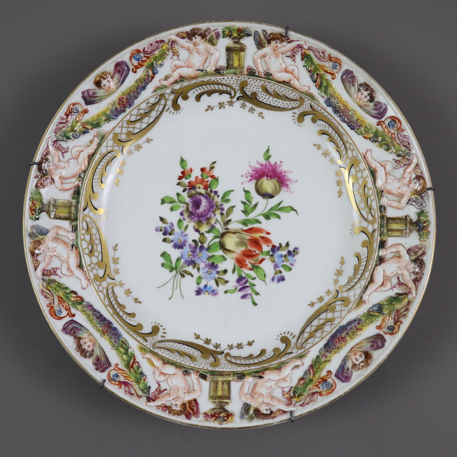 Zierteller mit Capodimonte-Reliefdekor - Neapelmarke, aber wohl Thüringen um 1900/Anfang 20.: Zierteller mit Capodimonte-Reliefdekor - Neapelmarke, aber wohl Thüringen um 1900/Anfang 20. Jh., Porzellan, Rundform, Fahne reliefiert mit Puttenpaaren Blumengirlanden haltend, polychrom bemalt,