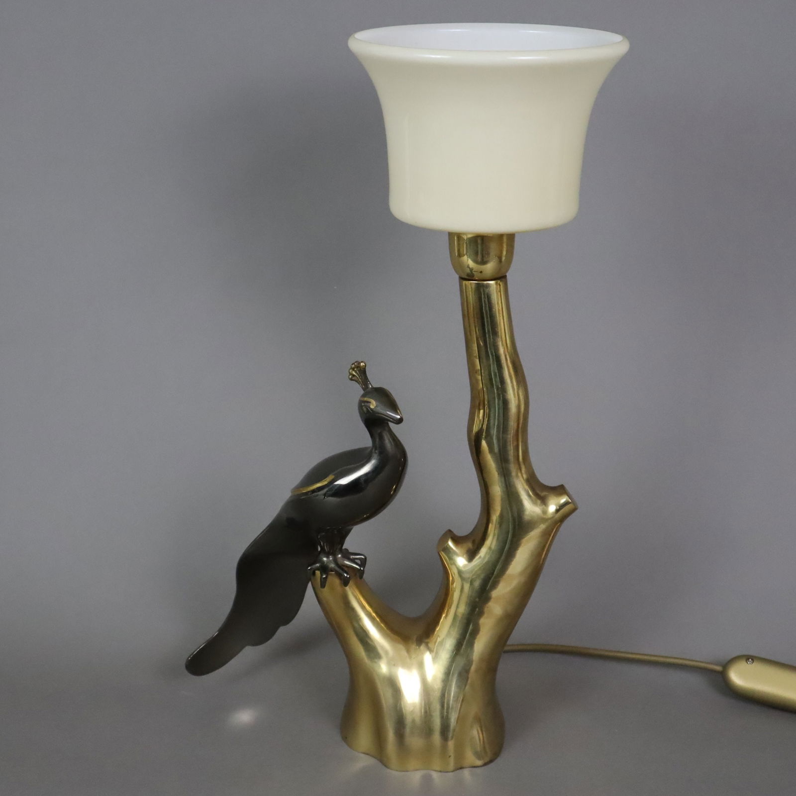 Tischlampe im Hollywood Regency-Stil - wohl Willy Daro, Belgien, 1960/70er Jahre, Messingfuß in: Tischlampe im Hollywood Regency-Stil - wohl Willy Daro, Belgien, 1960/70er Jahre, Messingfuß in Astform, darauf ein vollrund gearbeiteter Pfau sitzend, teils silberfarben patiniert, opalweiß