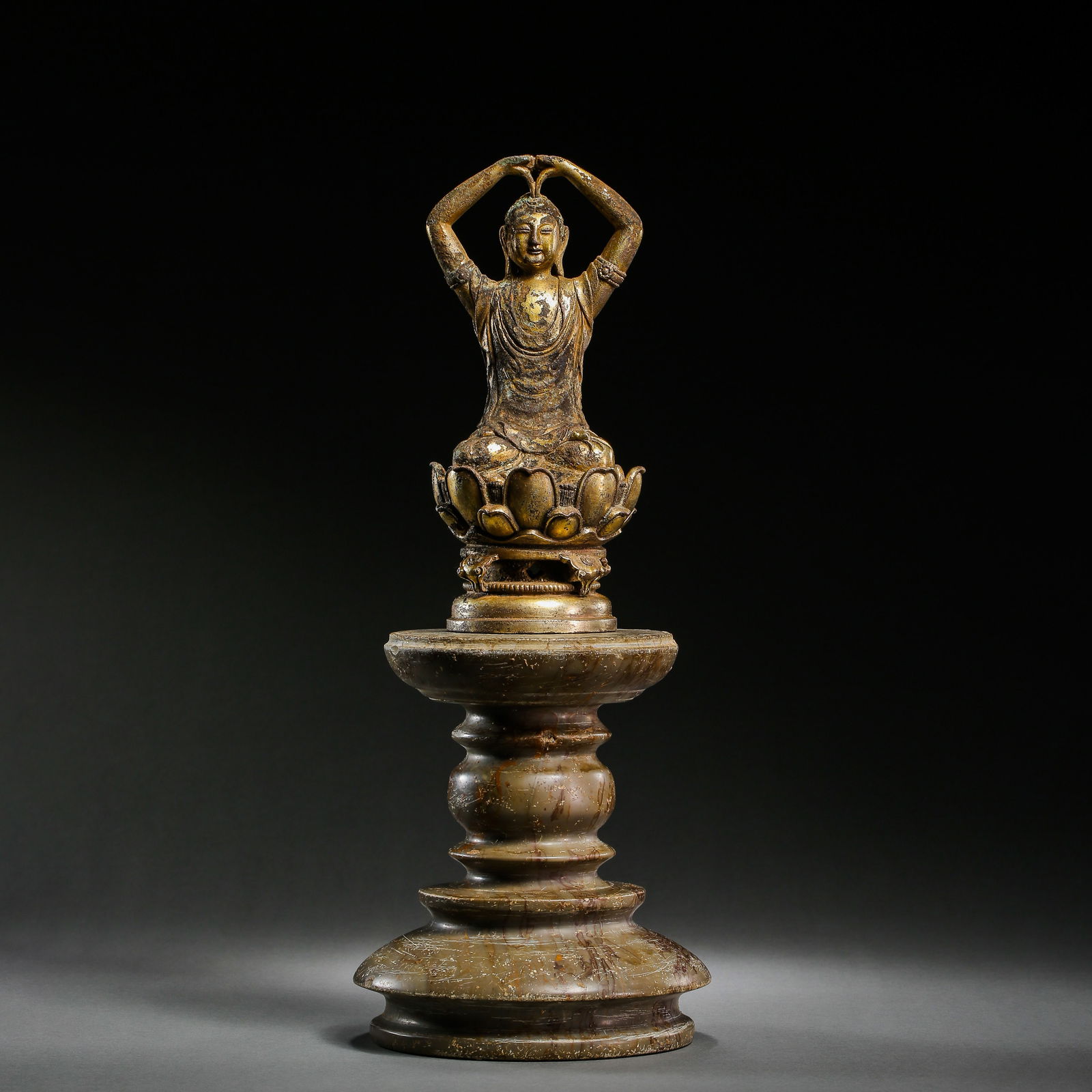 A RARE GILT-BRONZE FIGURE OF A SEATED BUDDHA ON A TIERED STONE PEDESTALLIAO DYNASTY (907-1125): The figure is shown seated in padmasana with hands raised above the head in an unusual mudra, the face with a serene expression and downcast eyes. The bronze retains significant traces of original gil