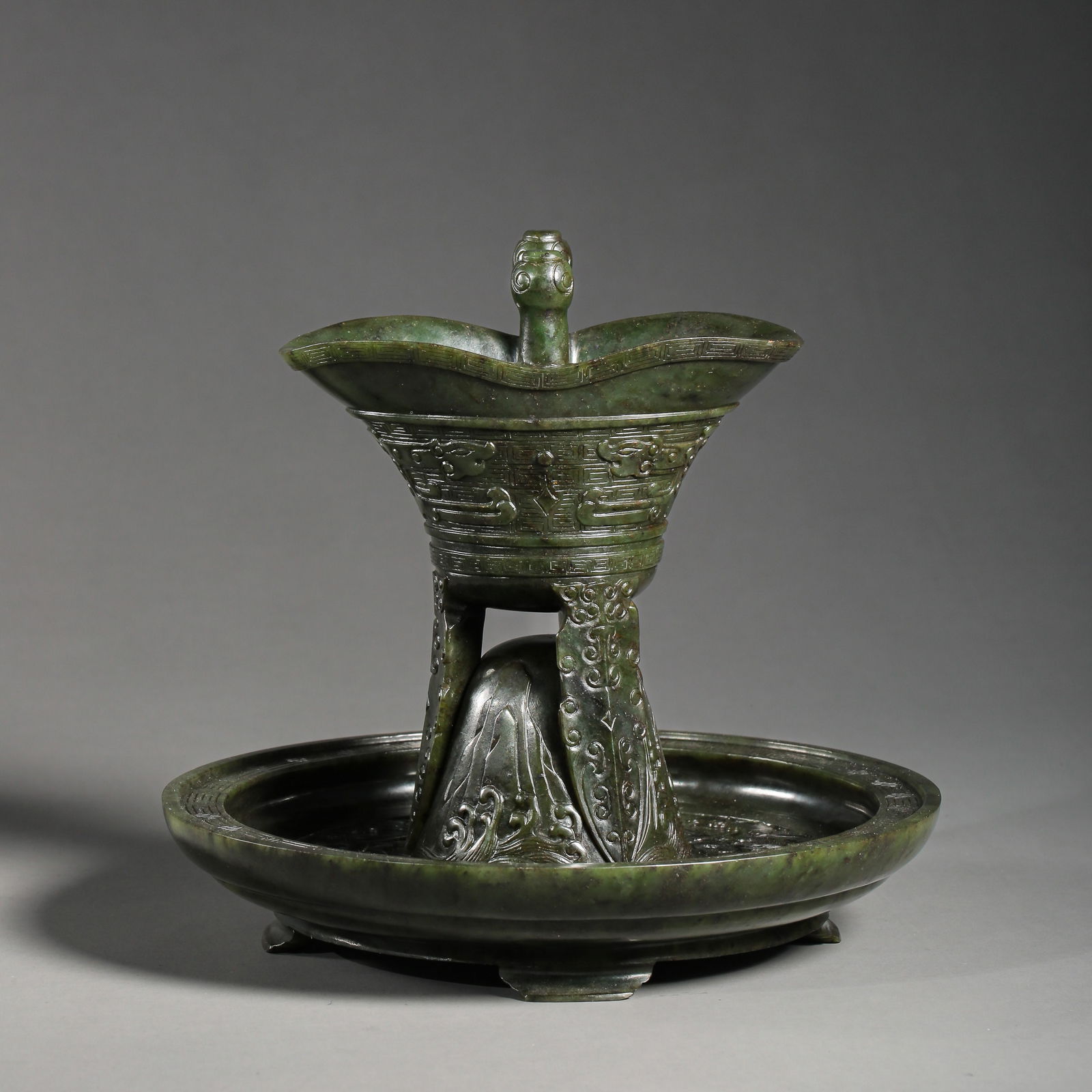 A RARE AND MAGNIFICENT SPINACH-GREEN JADE 'ARCHAISTIC' LIBATION CUP AND STANDMING - QING DYNASTY: The set is masterfully carved from an exceptionally rich, spinach-green jade (manqing) with subtle dark speckling characteristic of premium nephrite. The cup (jue) is modeled after ancient ritual bron