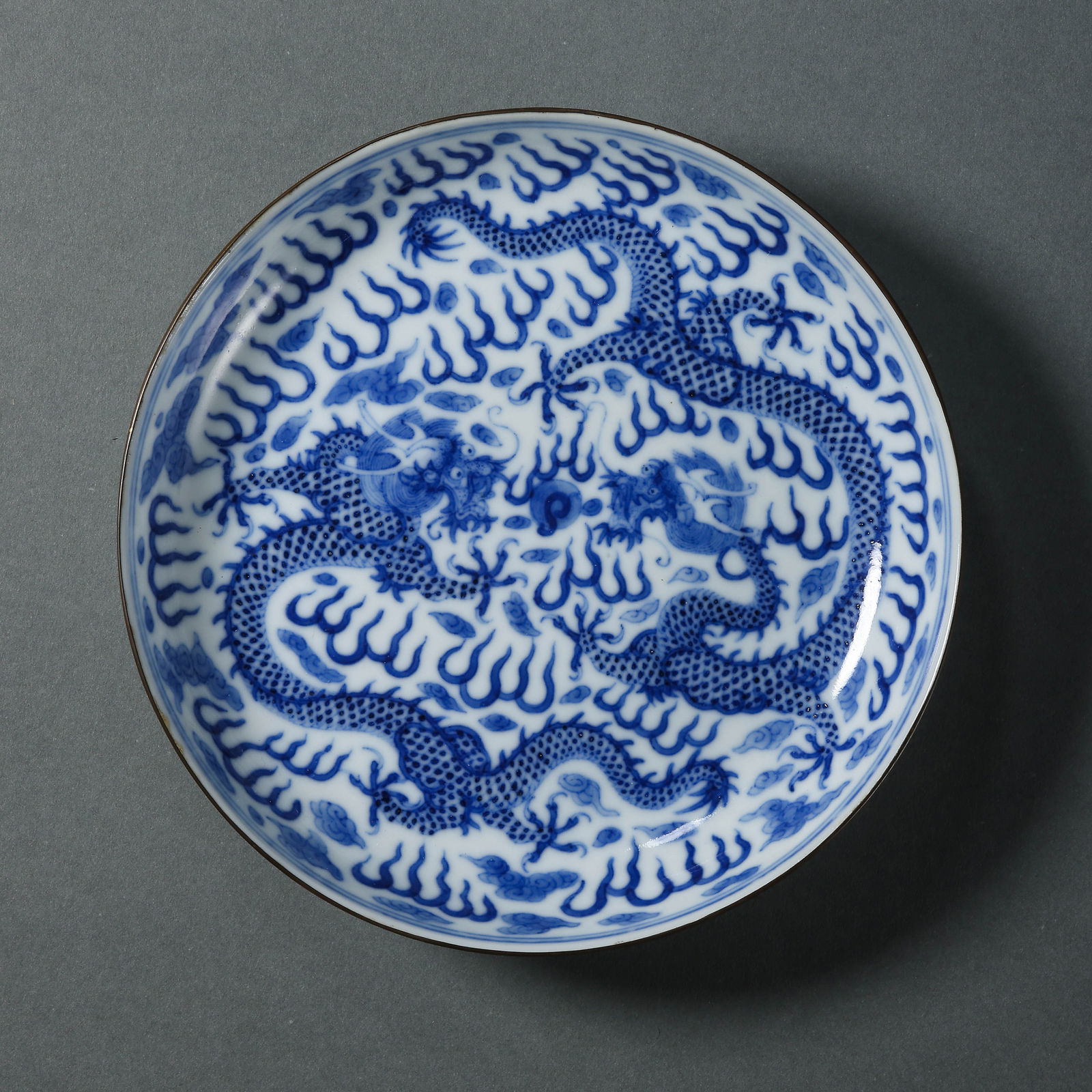 A BLUE AND WHITE 'TWO DRAGONS' DISH WITH METAL-BOUND RIMQING DYNASTY, 19TH CENTURY: Description The dish is of an elegant, shallow form with a subtly flared rim, which is meticulously metal-bound (bao kou) to protect the edge. The interior is vibrantly painted in underglaze blue with
