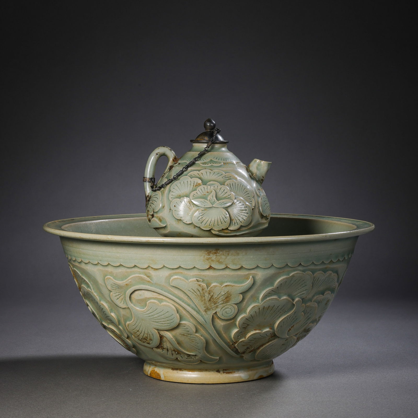 A RARE AND FINELY CARVED CELADON-GLAZED 'YUE' WARE EWER AND WARMING BASINFIVE DYNASTIES TO NORTHERN: DescriptionThe set comprises an elegantly proportioned ewer and a deep, matching warming basin .The Ewer: Featuring a multi-lobed, melon-shaped body, it rises to a flared neck and is flanked by a grac