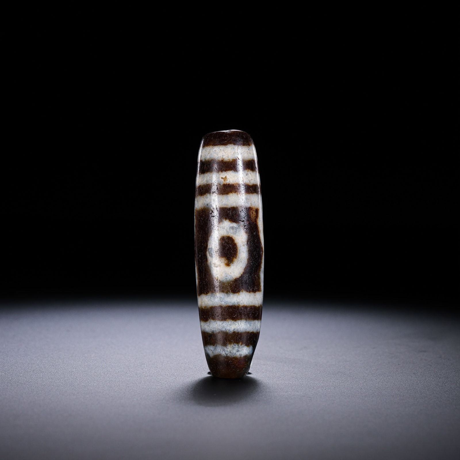 A RARE AND MAGNIFICENT MONUMENTAL 'THREE-EYED' DZI BEADHIMALAYAN REGION, TIBET, ANCIENT TO MEDIEVAL: Description This extraordinary Dzi bead is of exceptionally large size, masterfully fashioned from high-density silicate agate. It features a robust, elongated spindle-shaped profile that possesses a