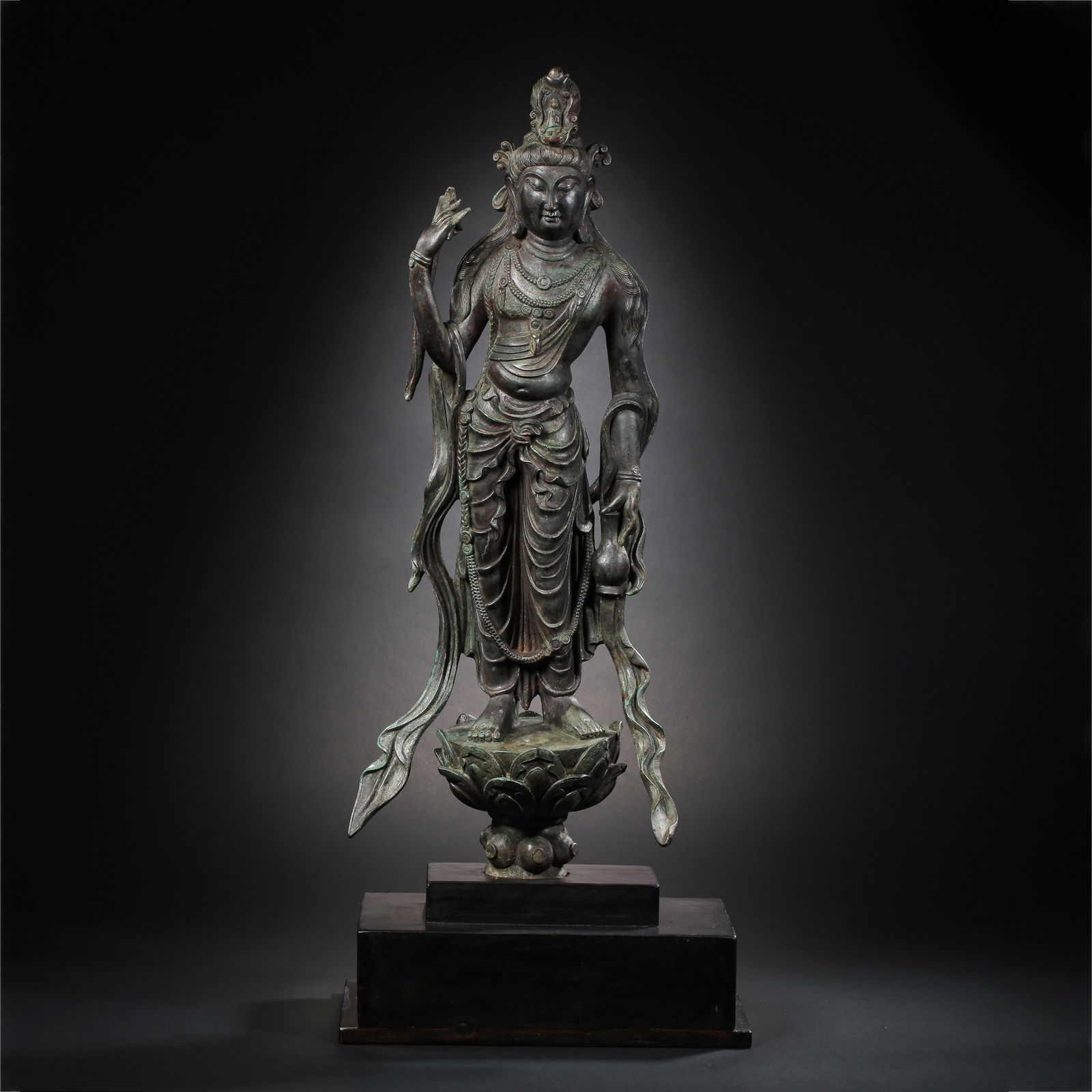 A RARE AND LARGE BRONZE FIGURE OF A STANDING GUANYINMING DYNASTY (1368-1644): DescriptionThe figure is cast with a serene and dignified expression, with downcast eyes and a gentle smile, flanked by long pendulous earlobes. The bodhisattva is elegantly draped in heavy robes that