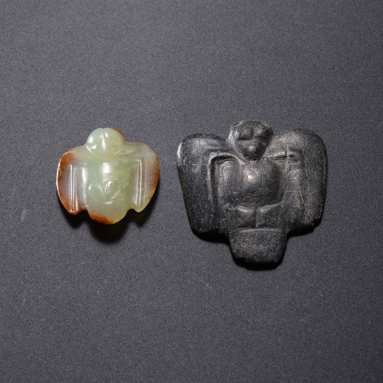 A RARE GROUP OF TWO HONGSHAN 'OWL' PENDANTSHONGSHAN CULTURE, NEOLITHIC PERIOD (C. 3500-3000 BC): Description The lot comprises two highly stylized bird-form pendants, identified as Owls (Xiao), each meticulously carved from contrasting Neolithic materials.The Jade Owl: Carved from a translucent g