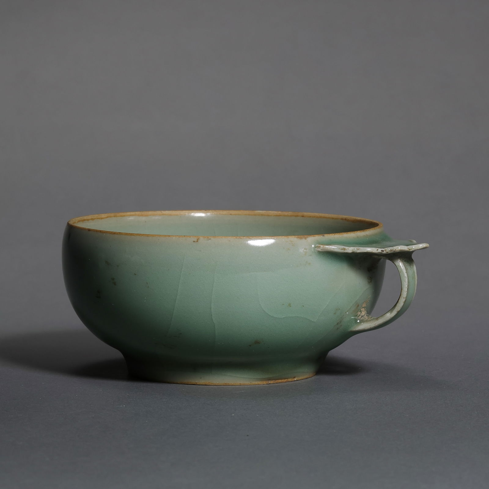 A RARE AND EXQUISITE LONGQUAN CELADON BOWL-FORM CUP WITH HANDLESONG DYNASTY (960–1279): Description This elegantly potted cup features a wide, flared body with shallow rounded sides, supported by a short, neatly trimmed foot. Attached to one side of the rim is a delicate, small loop hand
