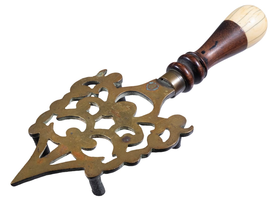 BRASS TRIVET WITH TURNED HANDLE: BRASS TRIVET WITH TURNED HANDLE Heavy Pierced Brass Trivet, having a turned wooden handle with carved ivory end cap, 11 3/4" long, 4 1/2" wide. OS: 11 3/4 x 4 1/2 in. (29.8 x 11.4 cm.)