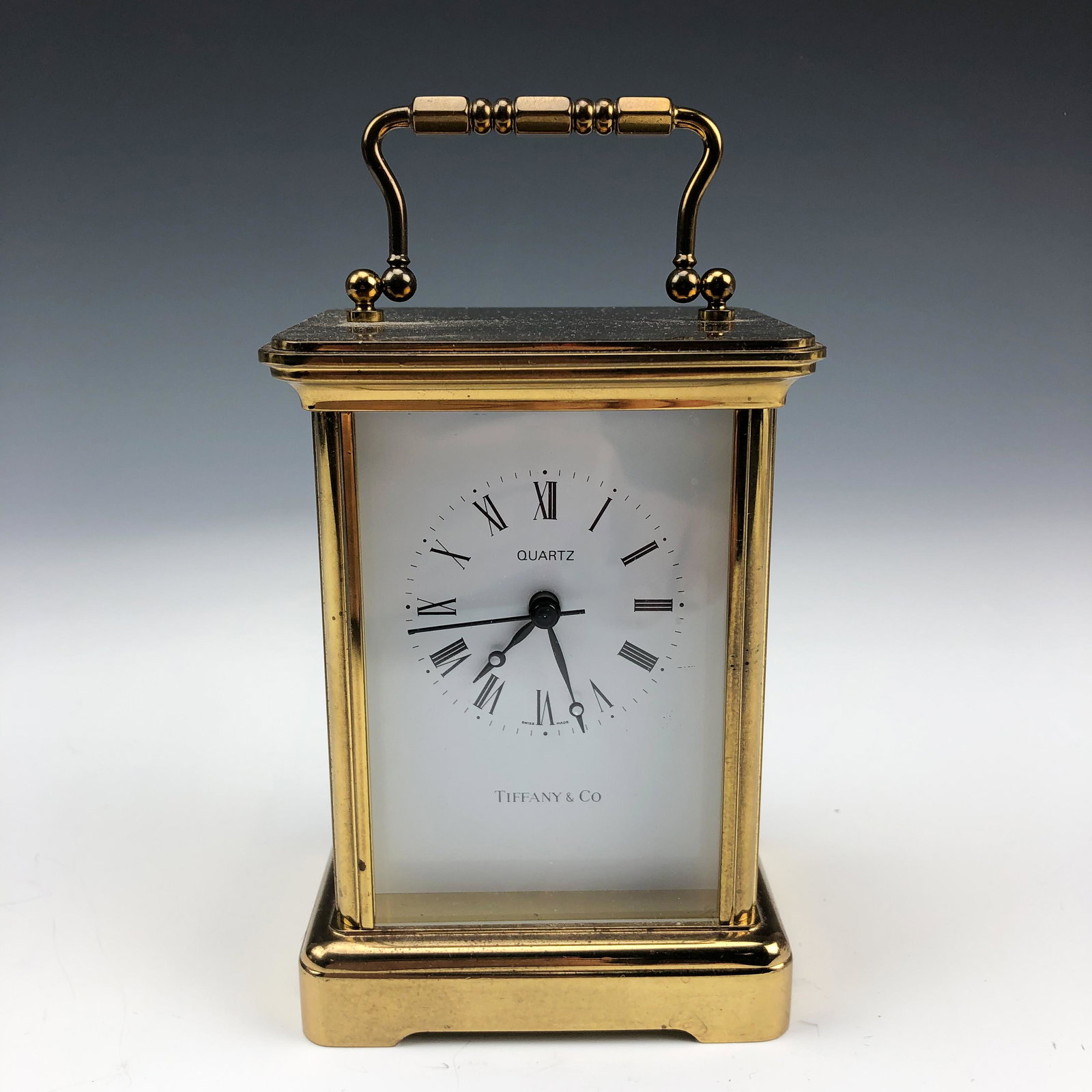 Tiffany Co Gilt Brass Quartz Swiss Carriage Clock: Features a Swiss Made Quartz Battery Operated Desk Carriage Clock. White dial face with bold Roman numeral markers. Black colored metal hands. Sturdy swivel handle atop. Polished finish to the case. M