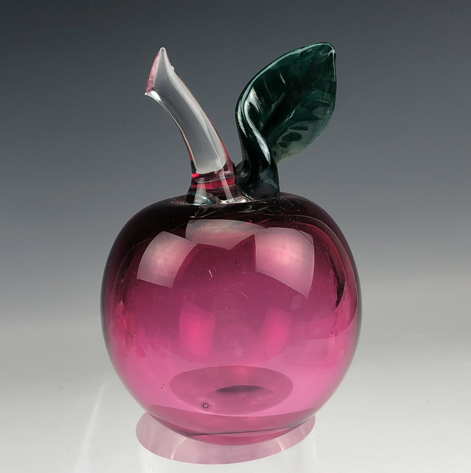 Mystery Artist Cranberry Glass Apple Paperweight: A painstakingly well crafted article of studio hand blown glass artistry with a warm nature inspired theme. Features a plump apple with an applied clear stalk and a green leaf. Smooth high mirror poli