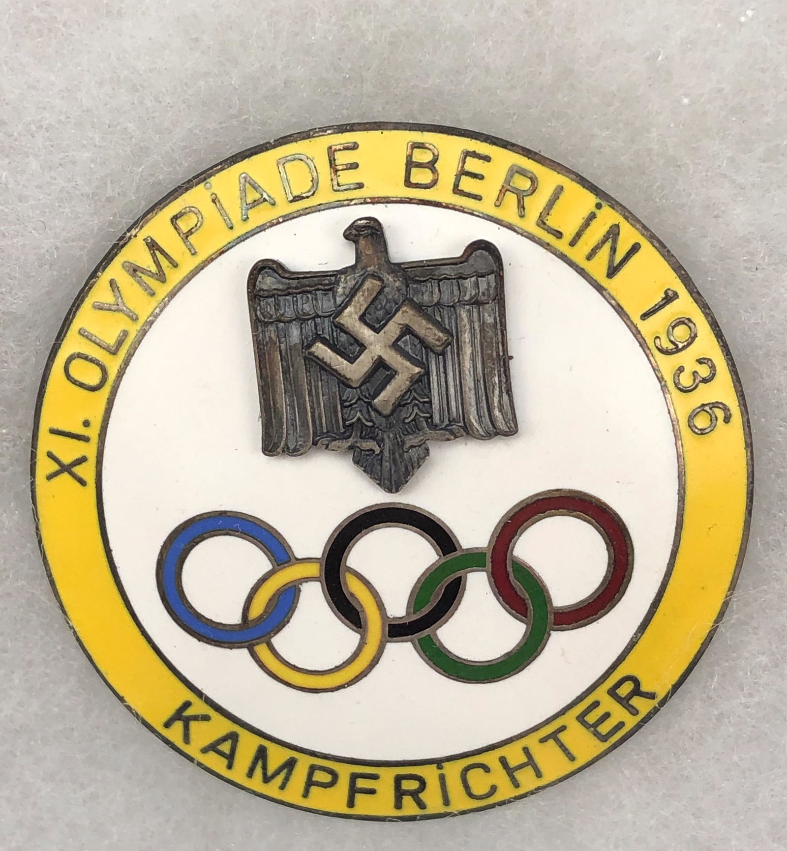 1936 Berlin Olympics Kampfrichter Judges Pin Badge: This 1936 Berlin Olympics Judges Badge designed with a white ground, the five colored rings and a yellow border. Warm original aged patina. Mounted in a glass pane box. Signed as shown in the picture.