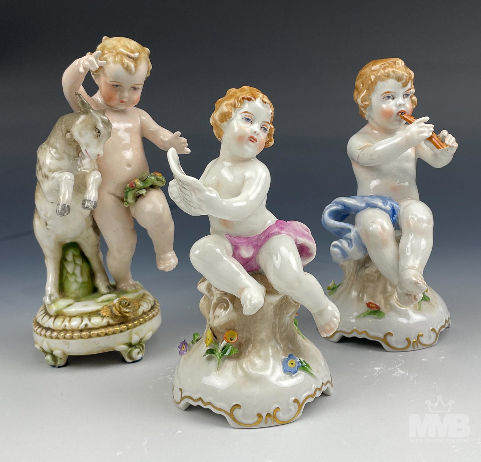 German Porcelain Nude Cherub Musician Figurine Lot: Three mythological heavenly nude angel cherubs, with one playing the flute, one holding a music score sheet and one frolicking with a baby goat. Warm hand painted colored with all having a glazed fini