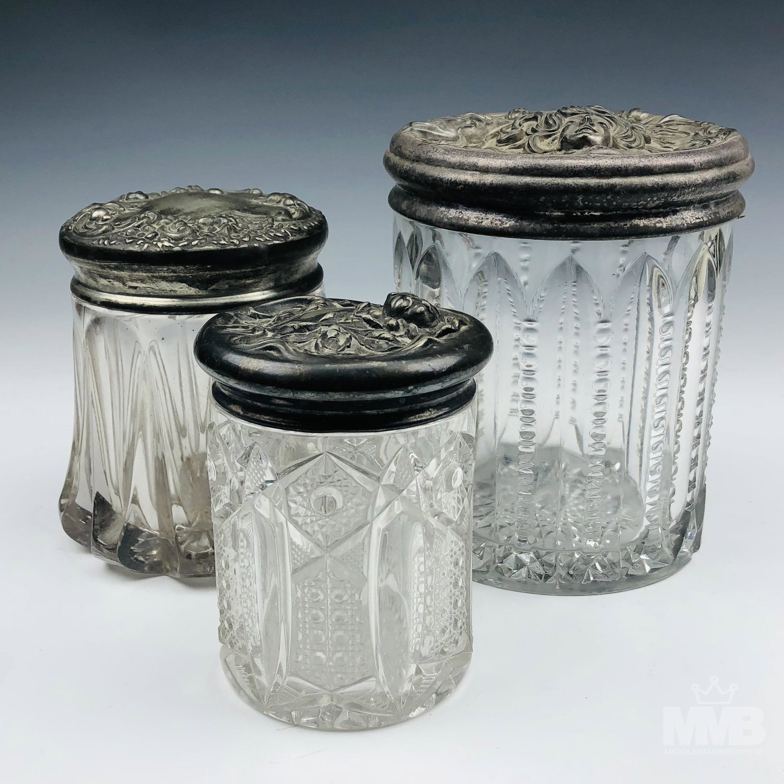 3 Antique Victorian Silverplate Glass Dresser Jars: Lid on each of the respective jar graced with a raised high relief Art Nouveau scroll and floral decoration, the largest one featuring a maiden's face with wavy flowing hair. The glass bodies feature 