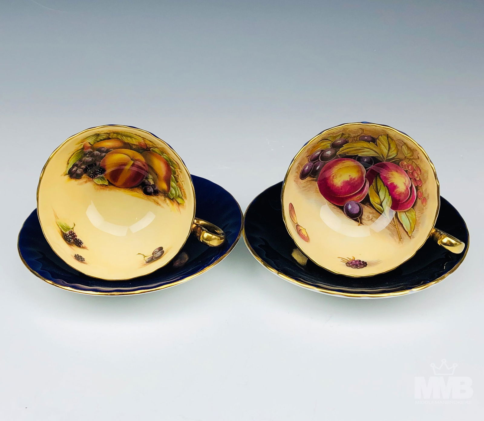 2 Aynsley Orchard Fruit Blue Tea Cup & Saucer Sets: Lot of 2 Aynsley fine English bone china cabinet display tea cups and saucers. Features cobalt blue with gold gilt trim and handle. Fruit design in interior of cup. Marked on bottom. Saucer measures .