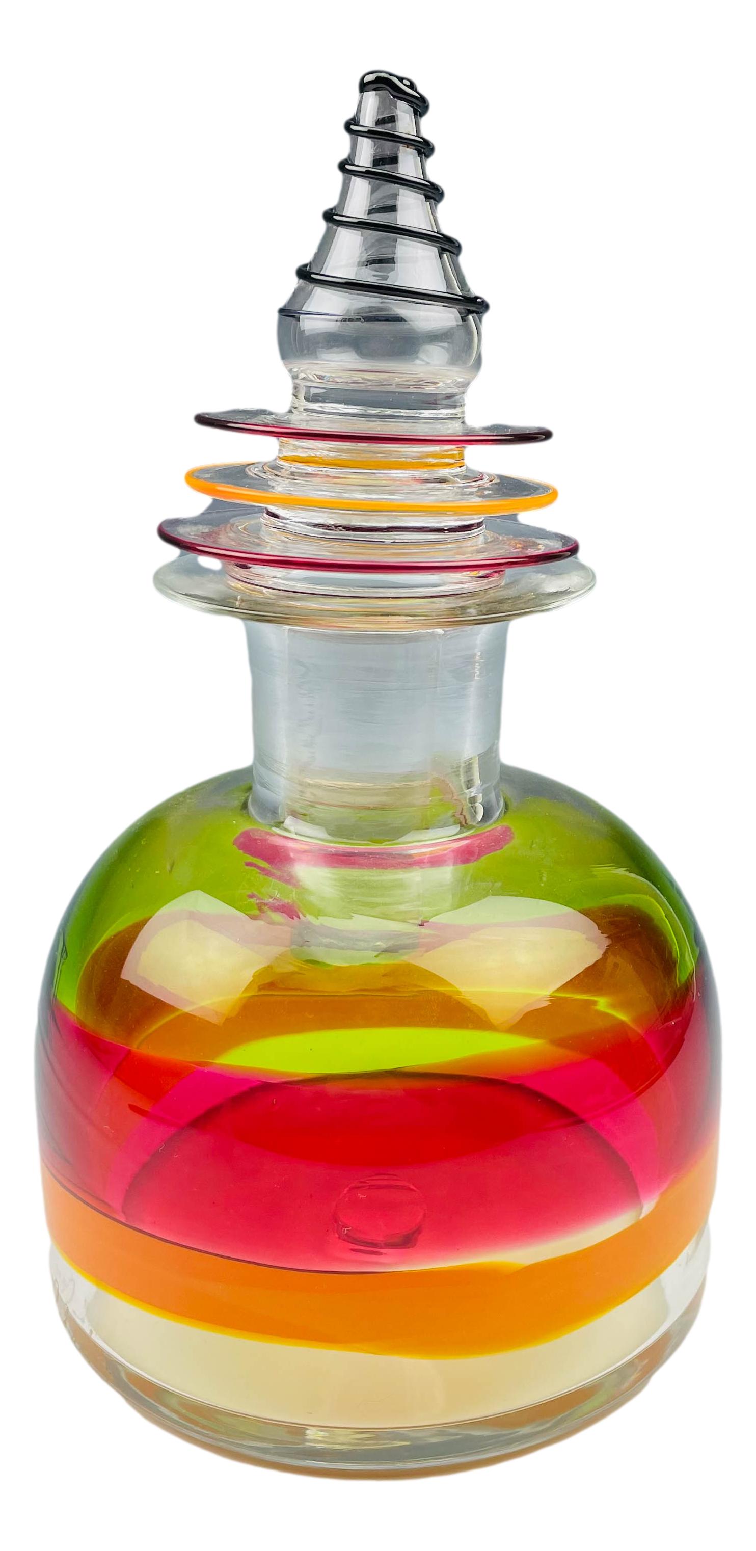 Modernist Studio Blown Cased Glass Liquor Decanter: Of simple shape with open mouth over short thick neck over low sloped shouldered body. A Multi Colored band encases the body around against a clear ground. Glass stopper with ringed design with applie