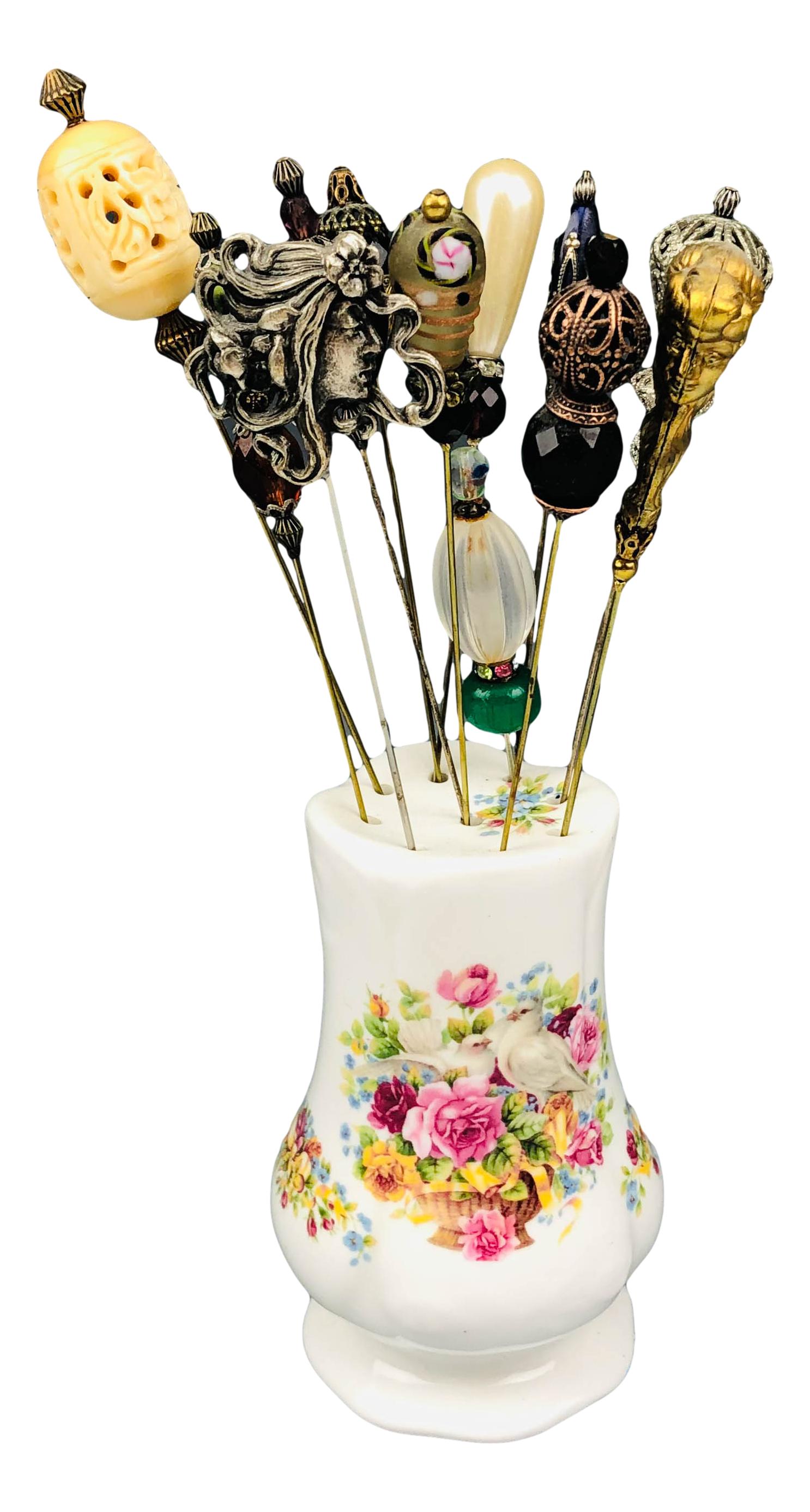 Collection Lot 12 Antique Stick Hat Pins w Holder: Includes a total of 12 stick hat pins, each embellished with a decorative top ranging from glass, Silver Plate, stone, faux pearl and natural elements. Accompanied in a porcelain holder with floral de
