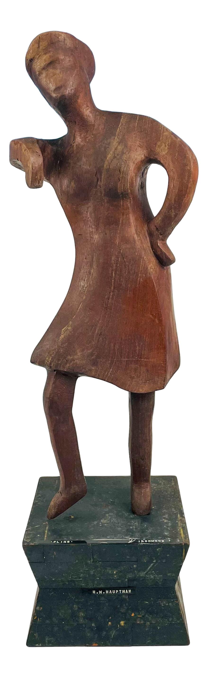 H W Hauptman Mid Century Figural Wood Sculpture: Subject matter features a woman in standing pose, one hand on her hip. Mounted atop a black colored geometric form wooden base. Measures 23 1/2". Signed ~ stamped on the black ticker reads "Fling" Iro