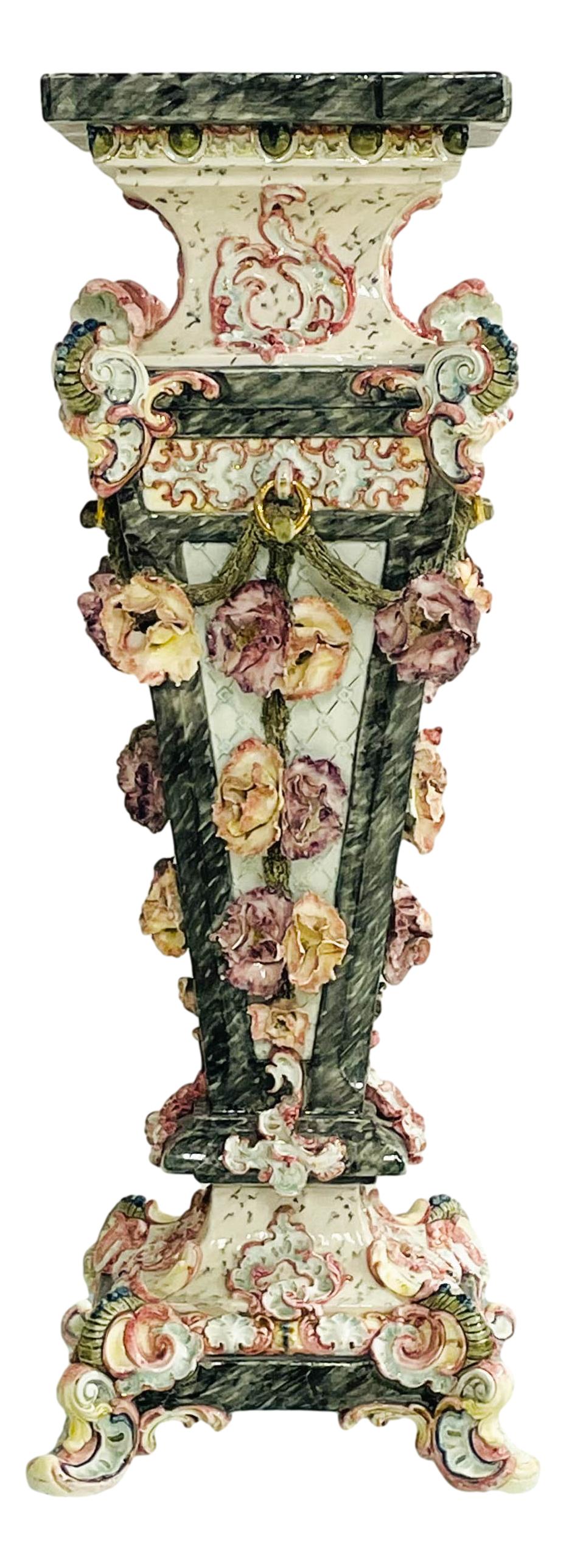 French Majolica Floral Porcelain Pedestal Stand: Majolica multi colored porcelain pedestal. Featuring raised relief pink and yellow floral design on a forest green ground. Dimensions: 39 X 12 X 12 in. Condition: Fair overall condition with mino
