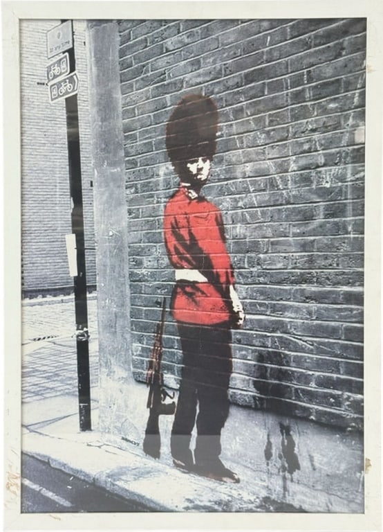 Framed Photographic Print in the Style of Banksy: Framed modern color photographic print on paper depicting London English street art in the style after Banksy. The image features a young Queens Guard in a red uniform against a brick wall, rifle slun
