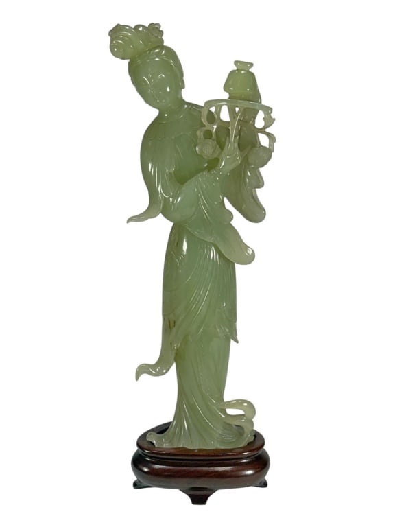 Chinese Carved Jadeite & Wood Figurine: Chinese jadeite figurine of a robed female holding a flowering branch and vessel. Displayed on a carved wood stand.Dimensions: 12.75 X 3.75 X 2.25 in. Condition: Good overall condition having norm