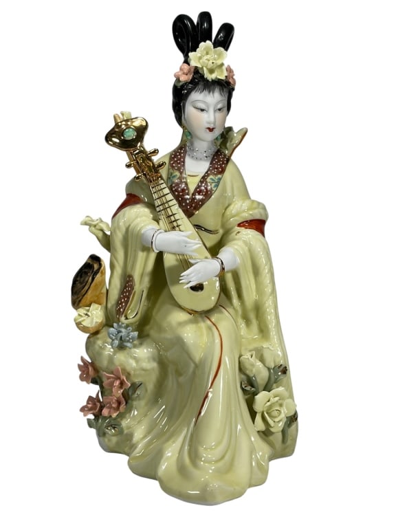Asian Geisha Playing Pipa Porcelain Figurine: Porcelain figurine of a seated geisha playing a pipa, decorated in yellow, red, and brown with applied flowers, gold highlights, and detailed hair ornaments.Dimensions: 15.75 X 7 X 7.5 in. Condition: