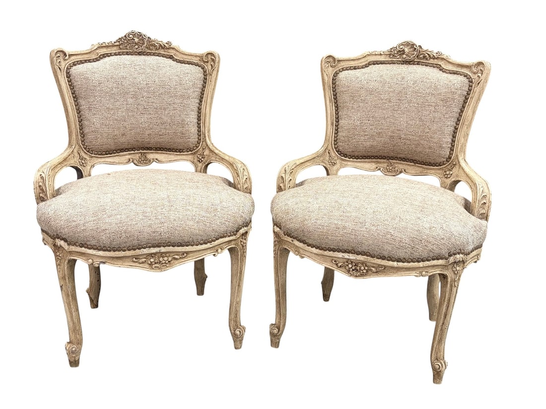 Pair of French Louis XV Style Beige Armchairs: Pair of French Louis XV style armchairs with carved floral and scroll frames, distressed painted finish. Beige textured upholstery with nailhead trim. Cabriole legs and curved arms. Each piece measure