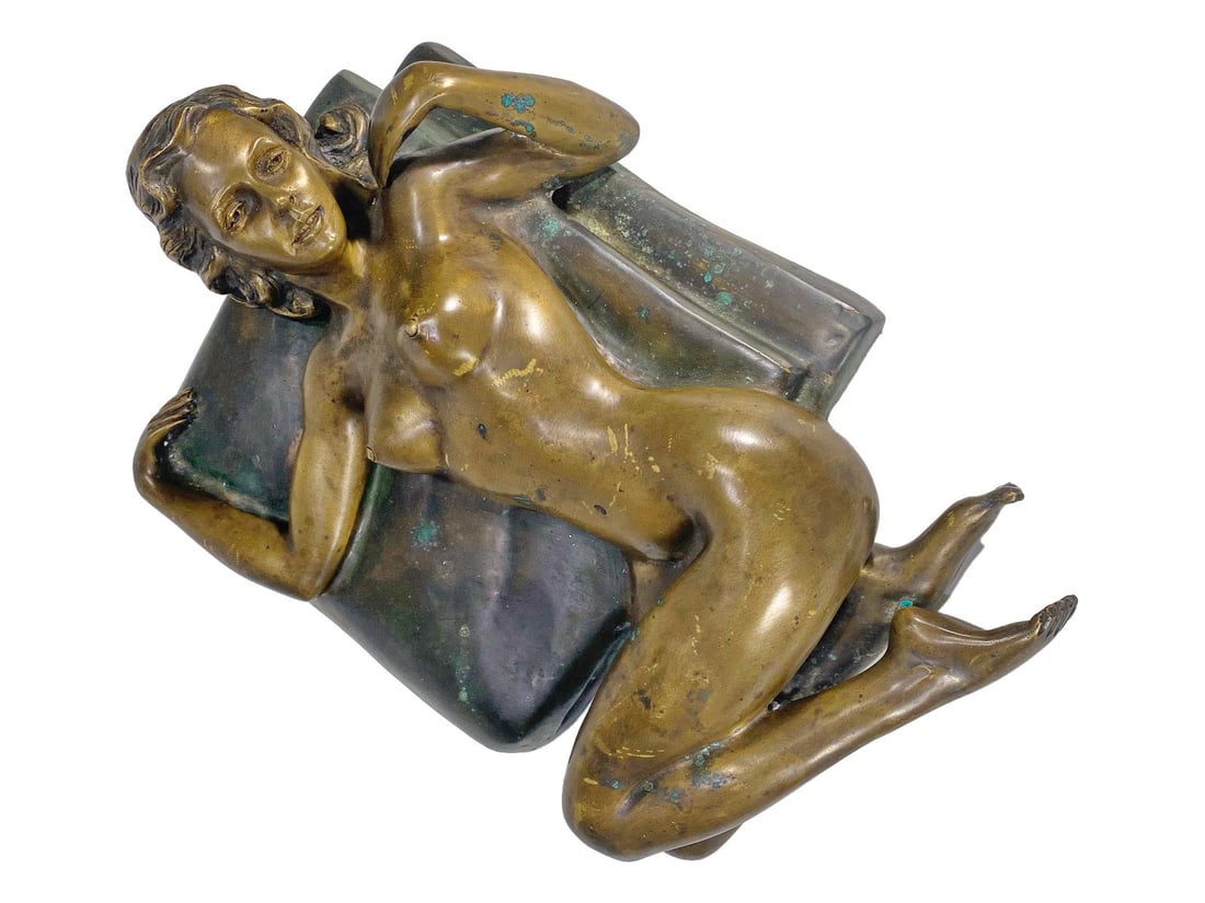 Reclining Nude Woman Figural Bronze Sculpture: Bronze sculpture of a reclining nude woman on a draped cushion, finely detailed and finished in warm patina with natural highlights.Dimensions: 7 X 10.75 X 15.25 in. Condition: Good overall condit