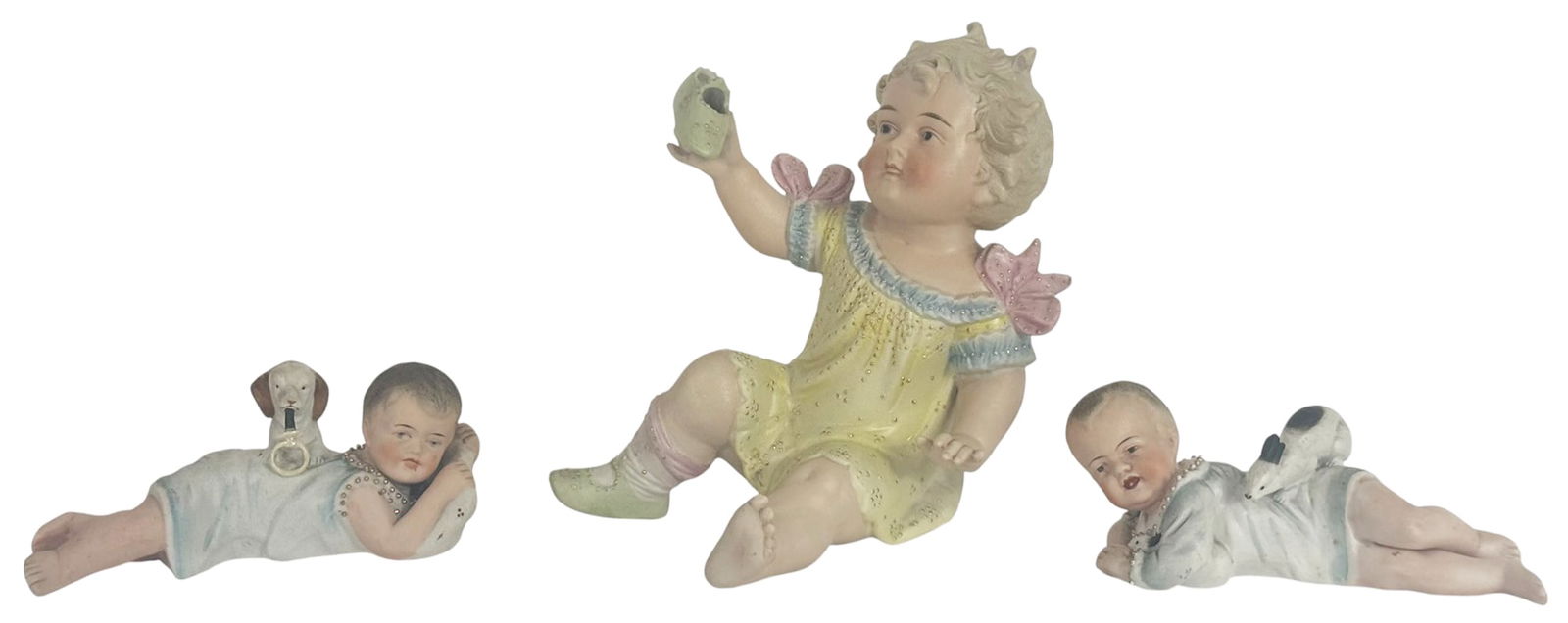 German Bisque Porcelain Baby Figurines 3pc LOT: Lot of three early 20th century German bisque porcelain baby figures. The set includes two reclining babies adorned in pale blue nightgowns, and the third of a seated child in a yellow dress holding a