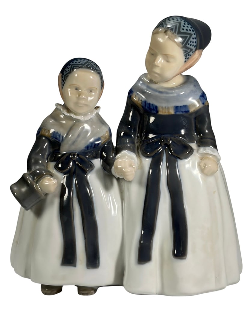 Royal Copenhagen Amager Girls Porcelain Figurine: Royal Copenhagen Denmark porcelain figure of two Amager girls in traditional dress. Both wear white skirts with dark blue bodices, long ribbon ties, and patterned headscarves, one holding a small pail