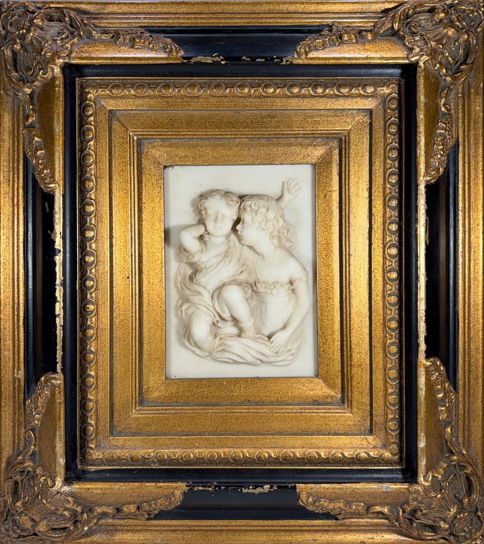 Framed Cherub Relief Plaque Composition Material: Framed cherub relief plaque, composition material, depicting two seated cherubs in high relief, set within a deep molded frame with gilt finish and black accent trim, featuring ornate corner embellish