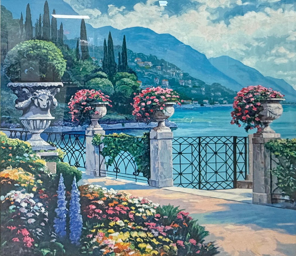 John Zaccheo Signed Limited Edition Coastal Print: Framed limited edition giclee print depicting a colorful lakeside or coastal garden terrace with flowering urns, wrought iron gates, cypress trees, mountains, and blue water beyond. Pencil signed by J