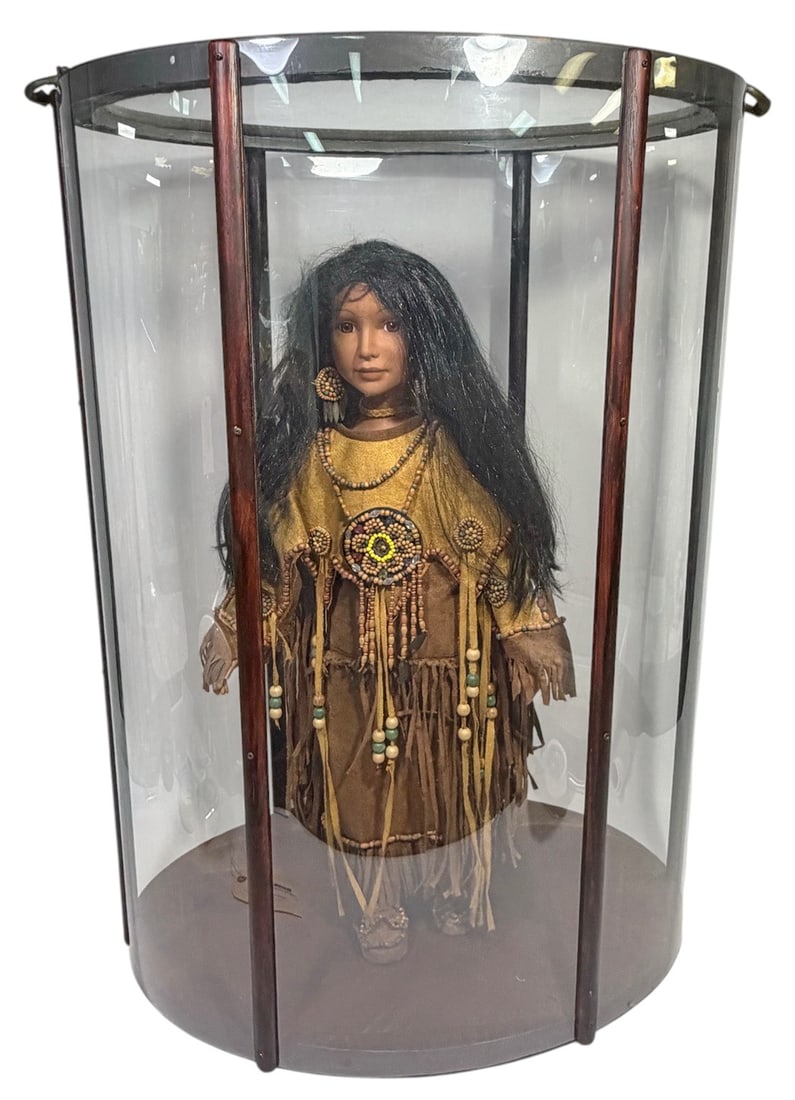 Native American Style Doll in Curved Display Case: Decorative doll dressed in Native American style attire with beaded ornaments and fringed garments, housed in a display case with wooden base, curved glass top, and clear plastic side panels. Features