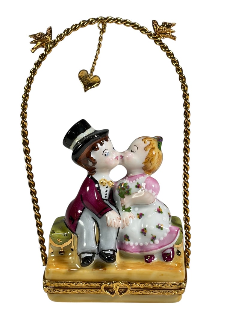 Limoges Bride & Groom Swing Trinket Box Sculpture: Hand-painted Limoges porcelain trinket box depicting a bride and groom seated together on a swing beneath a twisted gilt-tone arch. The couple share a kiss while a suspended heart charm hangs overhead