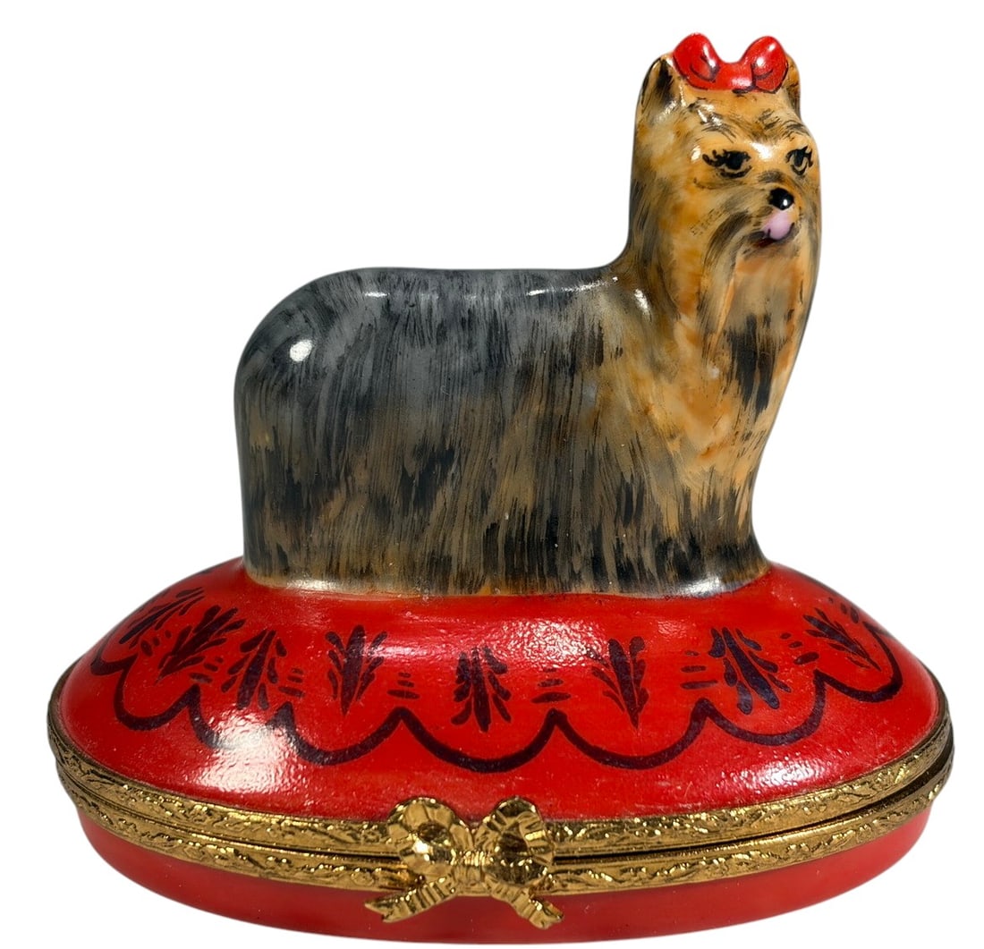 Artoria Limoges Yorkshire Terrier Trinket Box: Hand-painted Limoges porcelain trinket box modeled as a Yorkshire Terrier dog standing atop an oval red cushion base. Decorated with detailed fur coloration, red bow, and black floral accents on the c