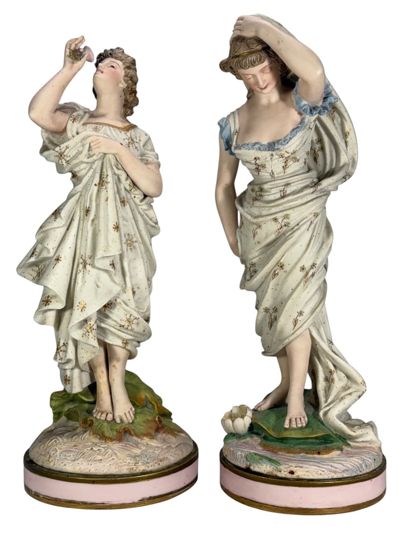 Pair Colored Bisque Porcelain Young Maiden Figures: Pair of colored bisque porcelain figures depicting young maidens in flowing classical attire. One figure raises a flower toward her face while the other adjusts a draped veil. Both feature hand-painte
