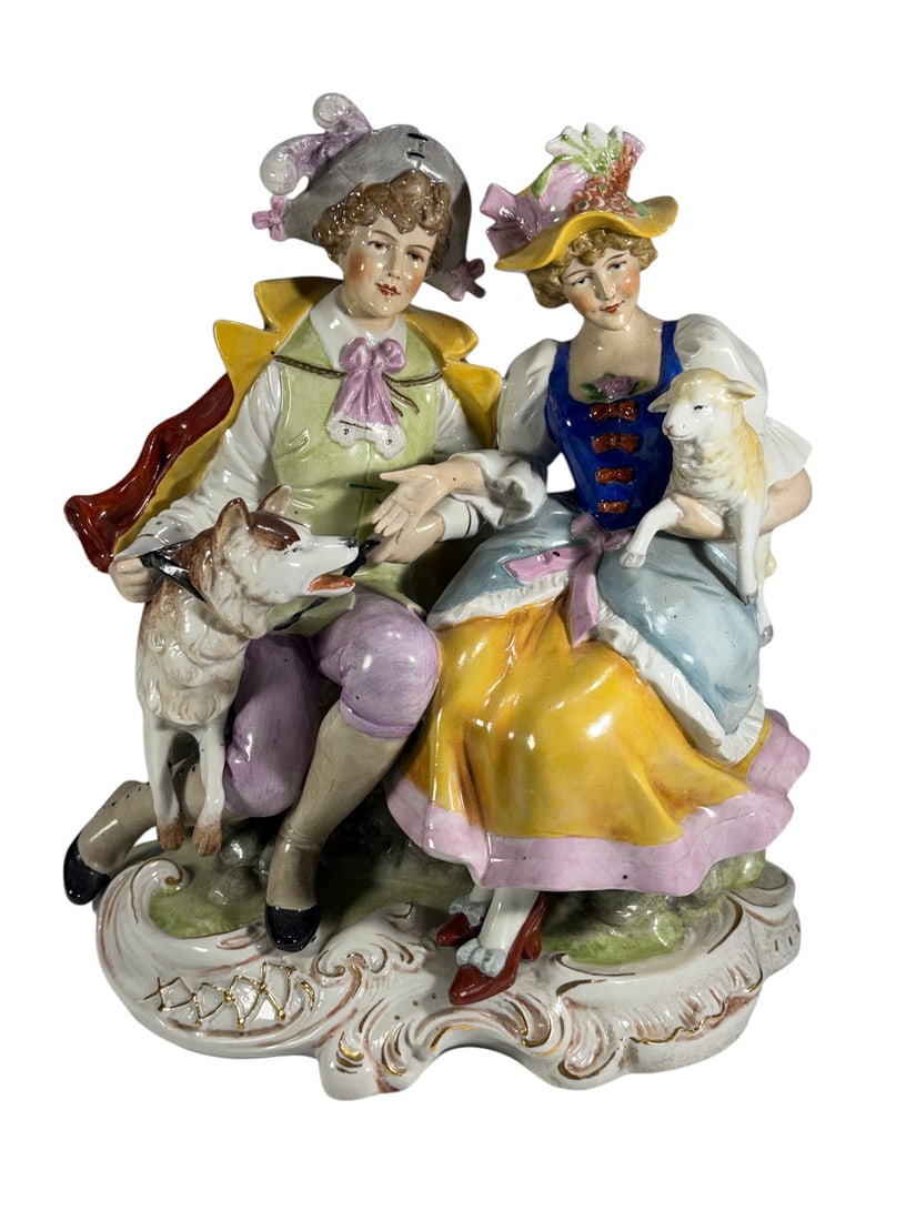 Paul's Gifts Germany Porcelain Couple Figurine Set: Colored porcelain figural group depicting an elegantly dressed young couple seated together on a naturalistic base. The gentleman is shown with a dog at his side, while the lady holds a lamb in her la