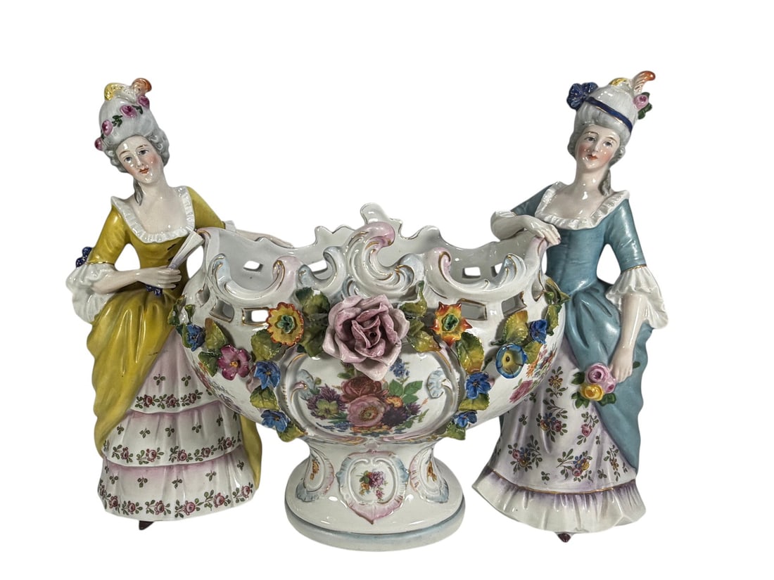 German Porcelain Figural Centerpiece Bowl Floral: German porcelain figural centerpiece bowl featuring two elegantly dressed ladies in period attire flanking a pierced Rococo-style bowl. Decorated with applied flowers, floral transfer panels, hand-pai