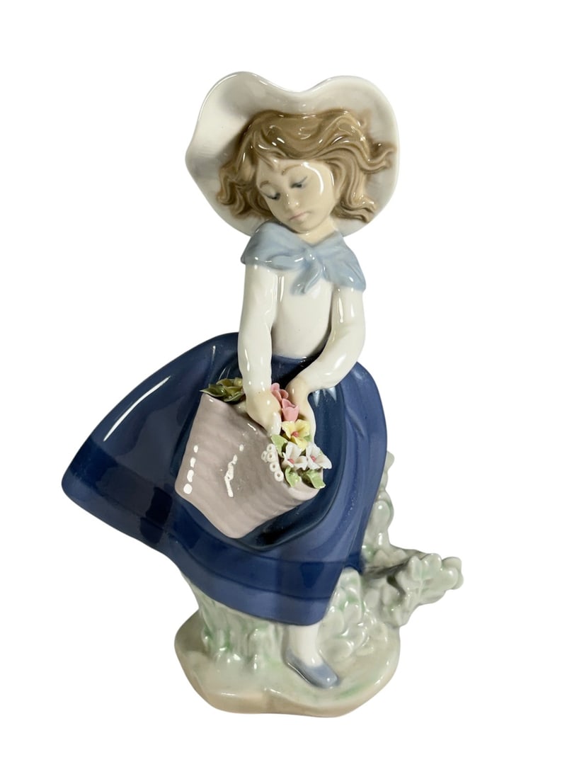 Lladro Spain Girl w/ Flower Basket Porcelain Fig: Lladro hand made porcelain figurine depicting a young girl wearing a wide-brimmed hat, blue dress, and neck scarf while holding a basket filled with applied flowers. The figure stands on a molded gras