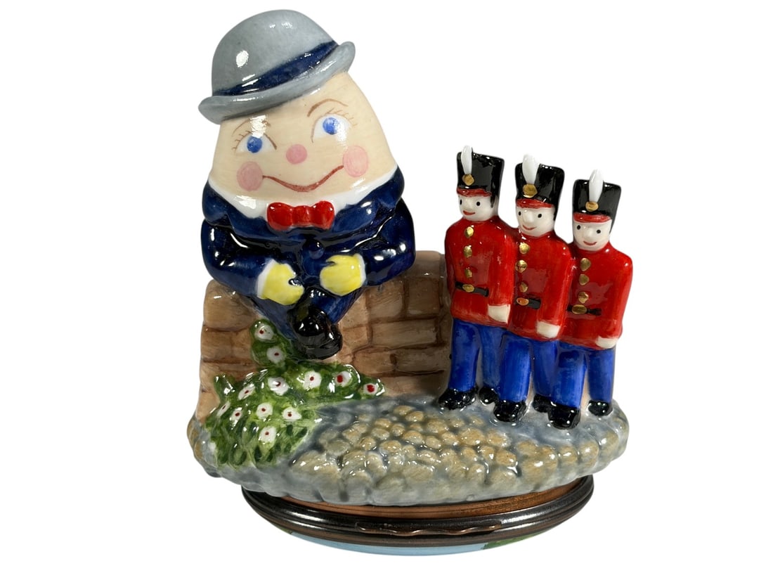 Humpty Dumpty Nursery Rhyme Trinket Box Figure: Porcelain figural trinket box depicting Humpty Dumpty seated atop a stone wall with three toy soldier figures standing nearby. The base is decorated with scenes and text from the classic nursery rhyme