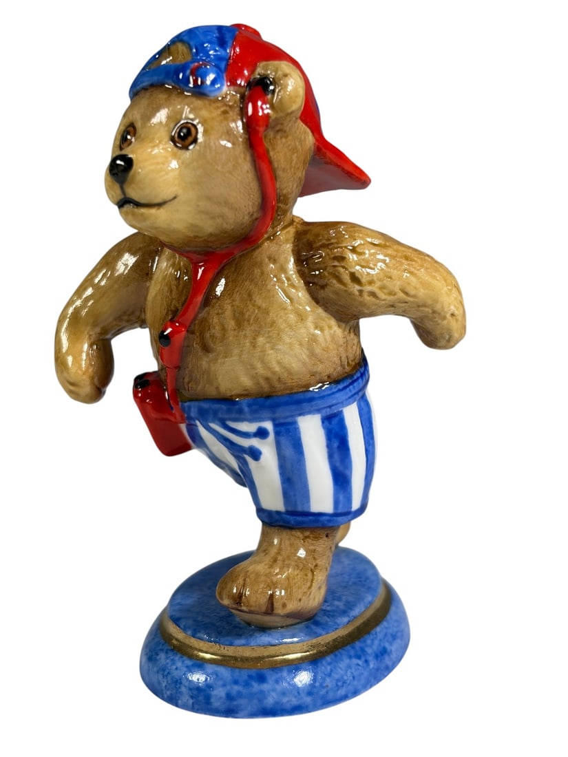 Halcyon Days Teddy Bear Porcelain Figurine Boxed: Halcyon Days porcelain teddy bear figurine depicting a bear dressed in blue and white striped swim trunks, red shoulder bag, and red and blue baseball cap. The figure stands on a blue circular base wi