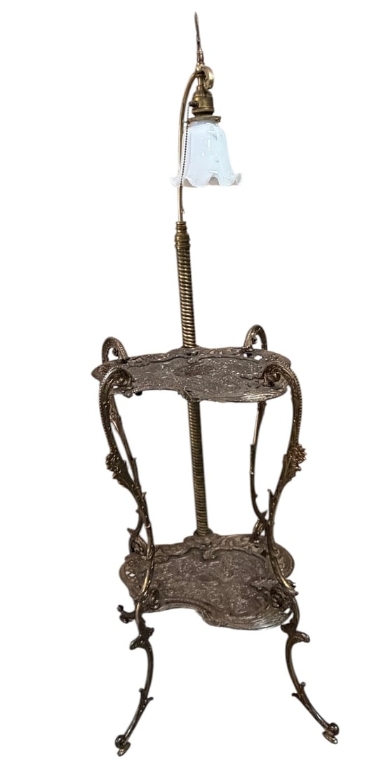 Victorian Style Brass Lamp Table & Magazine Rack: Ornate Victorian style brass-tone floor lamp table featuring two highly decorated embossed shelves, scrolling openwork supports, integrated magazine rack, and single light fixture with white glass sha