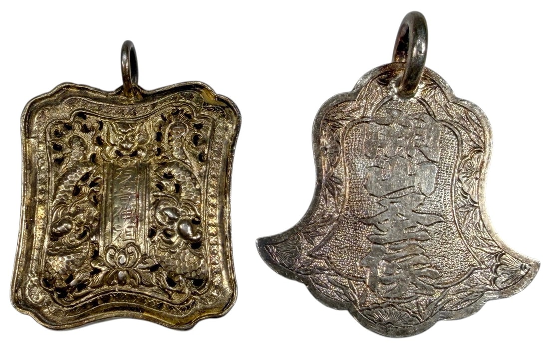 Pair of Chinese Low-Grade Silver Amulet Pendants: Pair of Chinese amulet pendants crafted in low-grade silver, each fitted with a suspension ring. One is rectangular with pierced and repousse dragon decoration surrounding a central inscribed panel; t