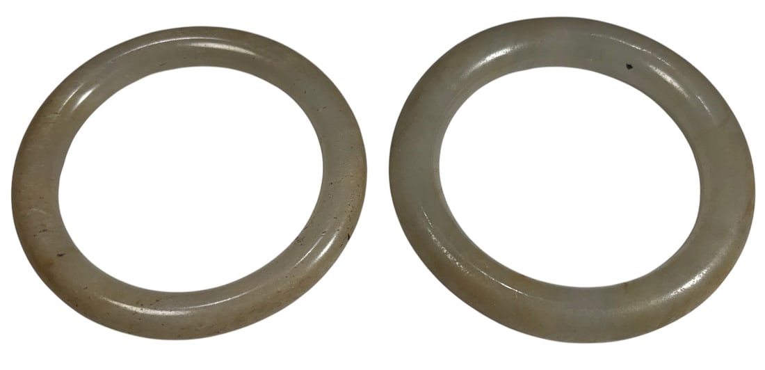 Chinese Pale Celadon-White Jade Bangles 2pc LOT: Pair of Chinese jade bangles in pale celadon-white tones, each of rounded circular form with smooth polished surfaces and natural color variations. One bangle shows a visible fissure or vein within