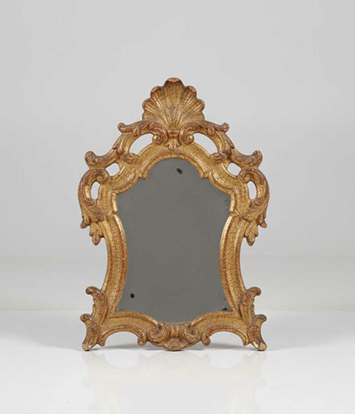 A Giltwood Rococo Style Mirror: Mirror set in a carved brass frame with a shell motif top. The decorative frame features scrollwork and a textured, worn finish. H: 34 cm
