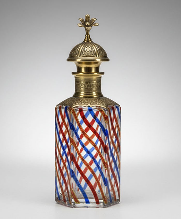 Gilt-Mounted Striped Glass Decanter: Glass bottle featuring diagonal red and blue stripes within the body. A decorative brass collar and a matching dome-shaped stopper with a floral finial sit on top. H: 14 CM