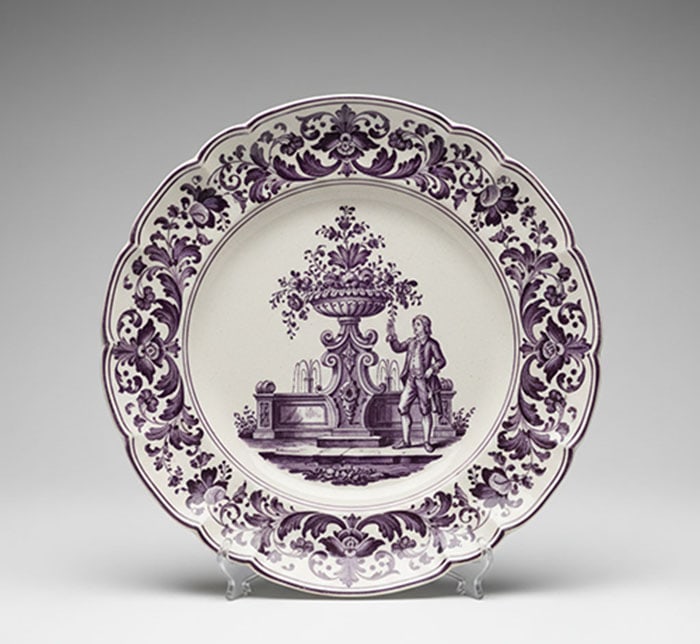 French Faience Manganese Glazed Plate: Circular ceramic plate features a scalloped edge and purple floral scrollwork around the rim. A central illustration depicts a man standing beside a large decorative urn and fountain. D: 15 CM