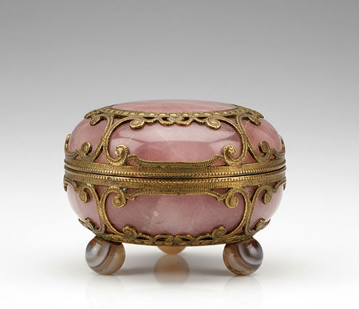 Gilt-Metal Trinket Box: Pink stone box featuring brass filigree accents around the lid and base. Small round banded stone feet support the structure from below. A thin brass rim outlines the opening of the lid. L: 14 CM