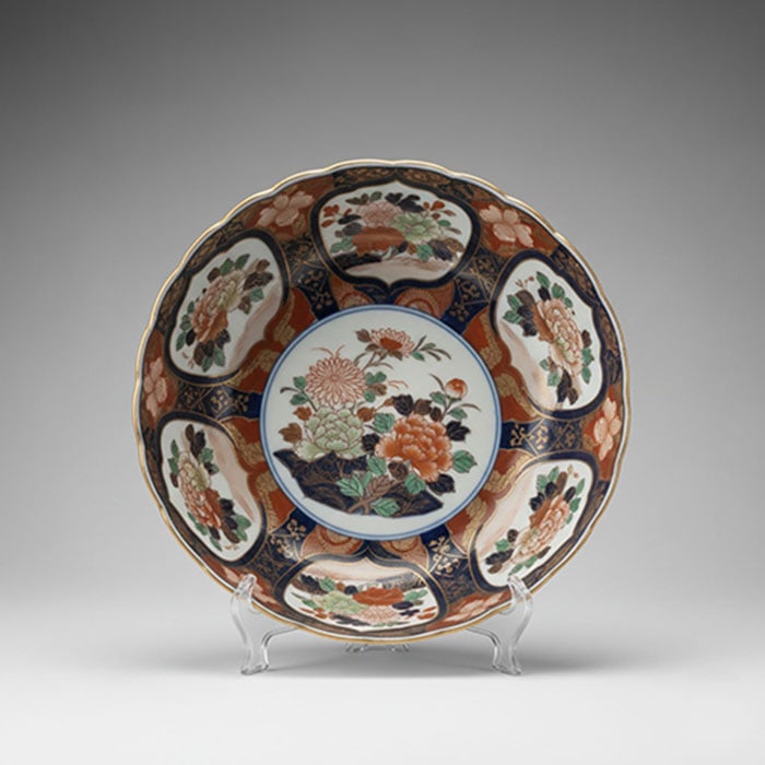 Large Arita Imari Porcelain Dish: Ceramic dish with a scalloped rim and a multi-colored floral design in orange, blue, green, and black. Brass accents are visible throughout the pattern, which features a central floral scene